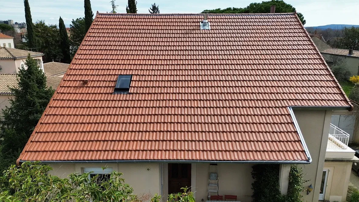 Toit rénové en Drôme à Montélimar avec des tuiles rouges modernes, présentant une fenêtre de toit et une cheminée, entouré de végétation et de bâtiments voisins