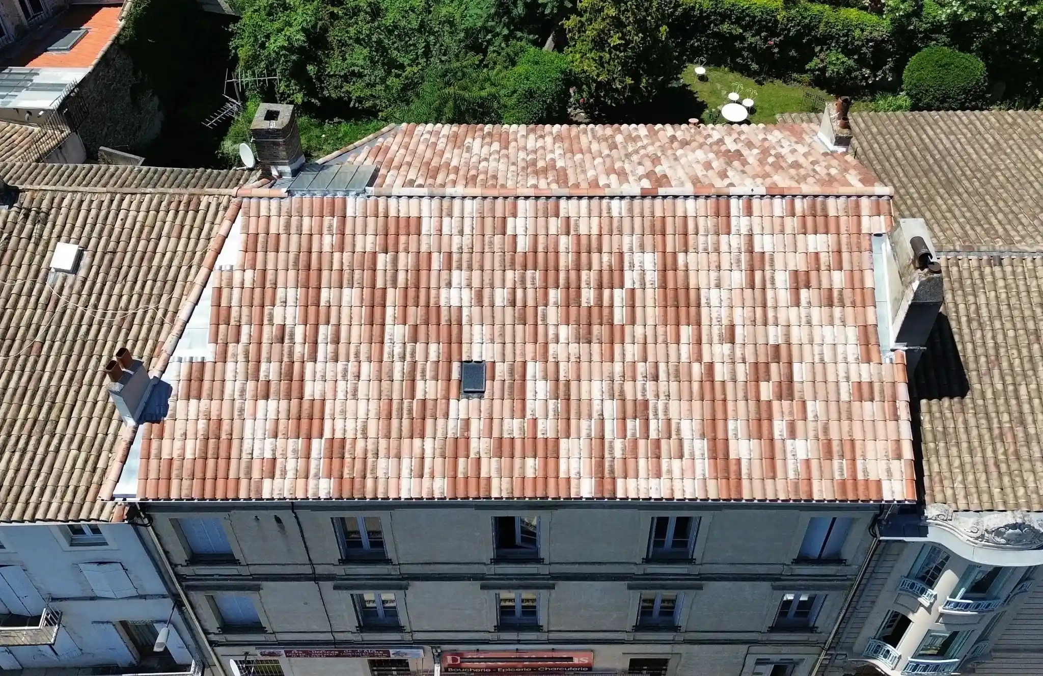 Toit en tuiles rouges et blanches d’un bâtiment résidentiel ancien, entouré d'autres toits et d'un jardin verdoyant en arrière-plan.