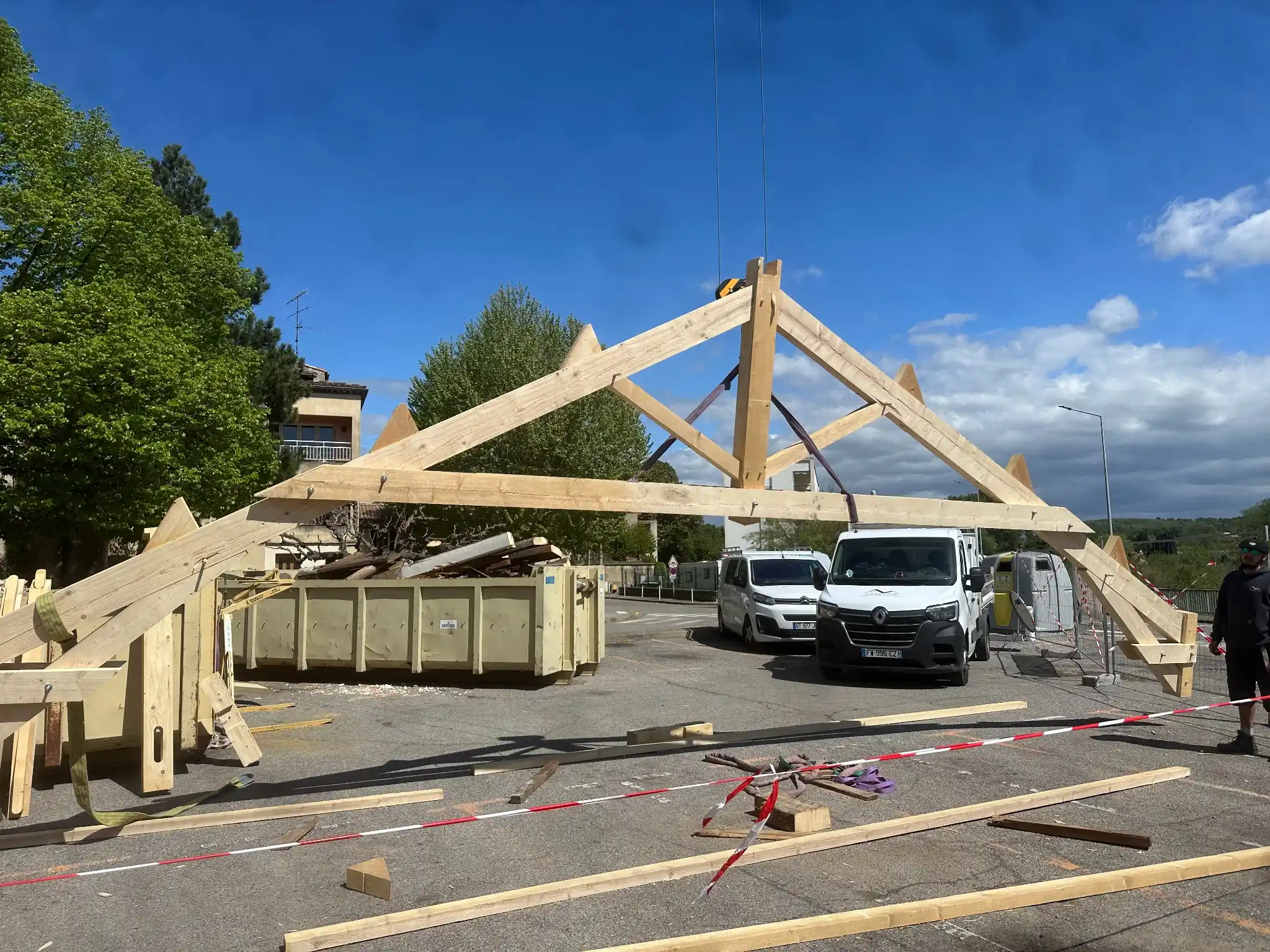 Grande structure en bois en cours d'assemblage sur un site extérieur, montrant les poutres en bois massives et des voitures en arrière-plan, partie du processus de construction de la toiture.