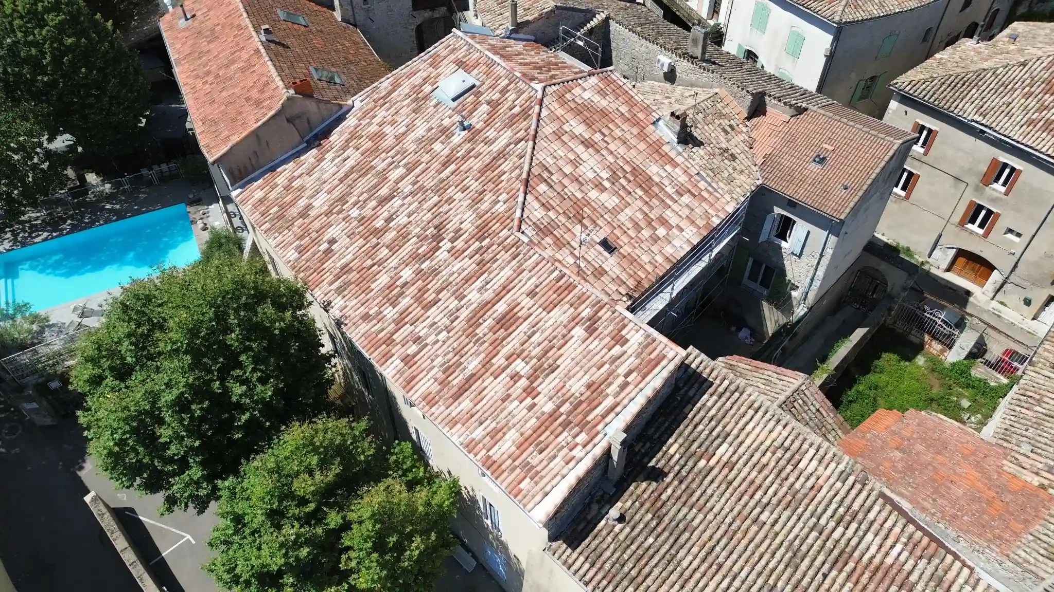 Vue aérienne d'un bâtiment avec un toit en tuiles rouges et blanches, entouré de bâtiments voisins et d'une piscine bleue à côté.