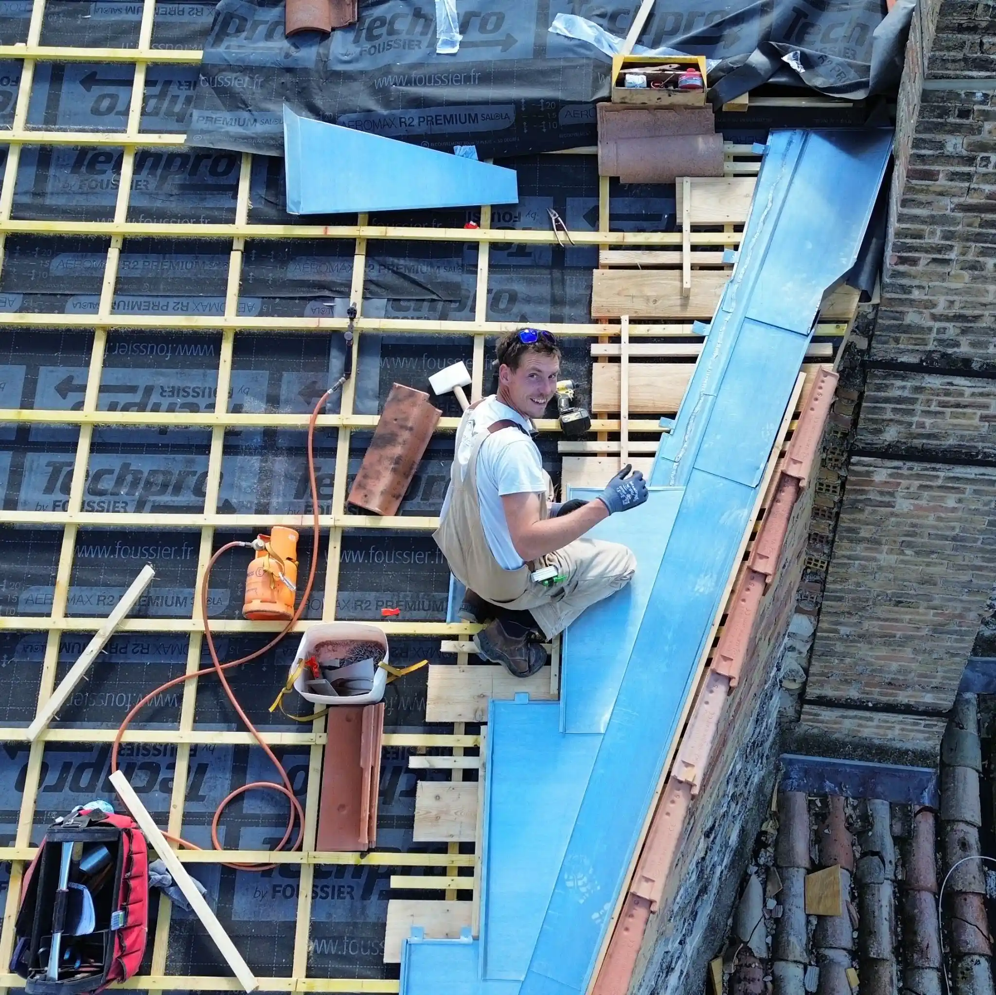 Ouvrier travaillant sur la structure du toit, entouré de matériaux de construction et de tuiles, en train de poser des éléments d'étanchéité